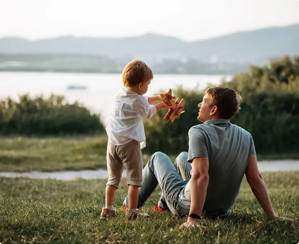 Son plays with wooden airplane next to his dad at a park